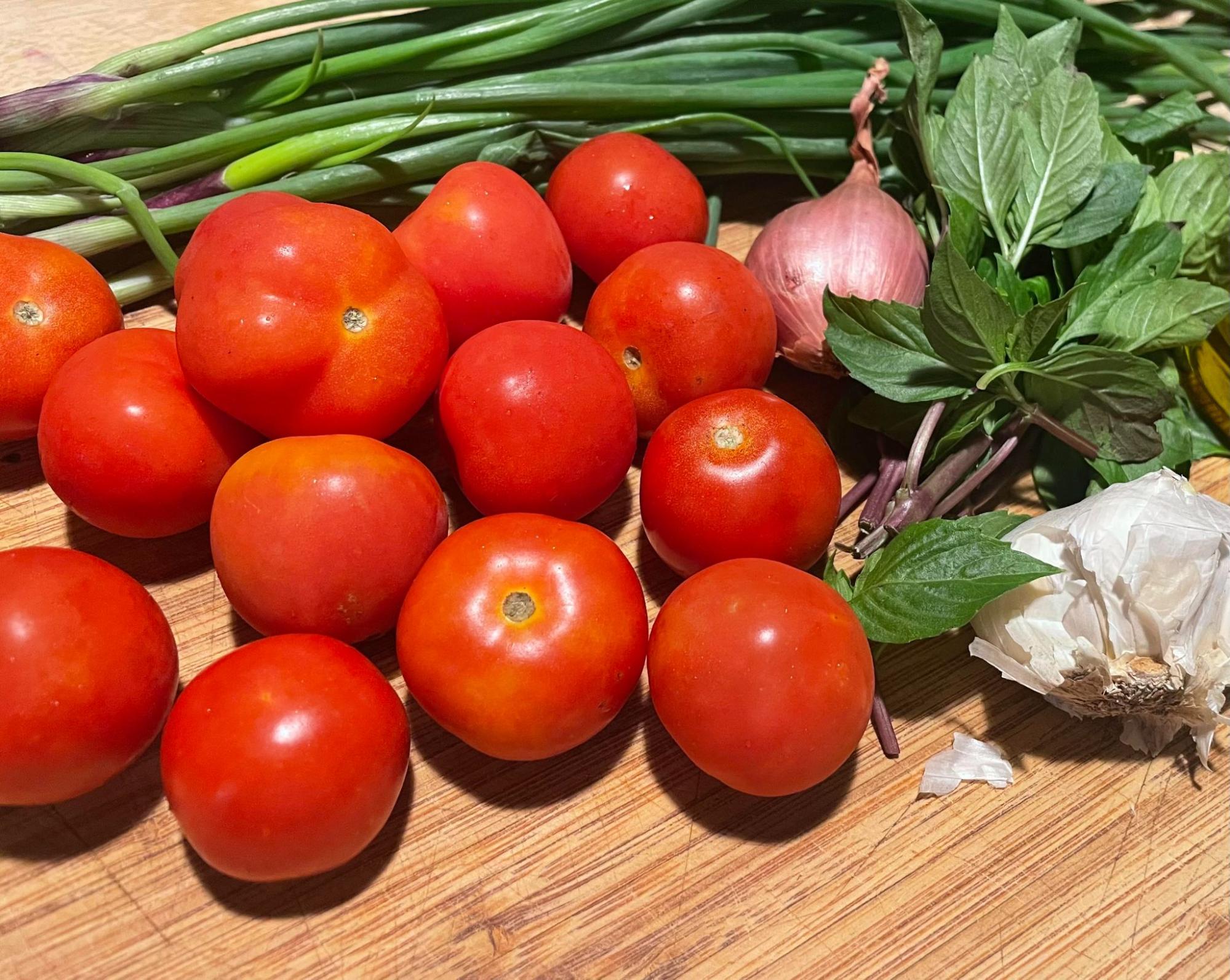 A cluster of bright red tomatoes on a tan cutting board with vibrant green basil leaves, a white head of garlic, green onions with specks of purple near their ends, and a light purplish scallion.